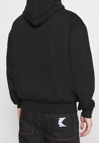Felpa nera con cappuccio regolabile, polsini e orlo a costine, abbinata a jeans scuri con un logo bianco sulla tasca posteriore.
