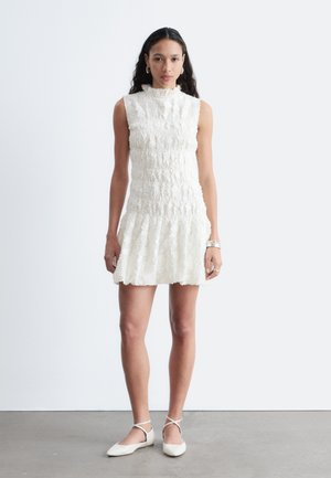 Žena stojaca, oblečená v bielej bezrukávovej štruktúrovanej šaty s volánovou lemom a bielymi balerínkami s členkovým remienkom na jednotnom pozadí.