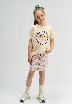 Cremfarbendes T-Shirt mit "Girl Power"-Grafik, kombiniert mit einem rosa, blumigen Knopf-Frontrock. Das Modell trägt weiße Sneaker und gestreifte Socken.