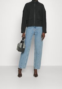 Bomber jacket nera con texture, chiusura con zip, due tasche frontali, abbinato a jeans a vita alta di un blu chiaro e una borsa a fantasia.