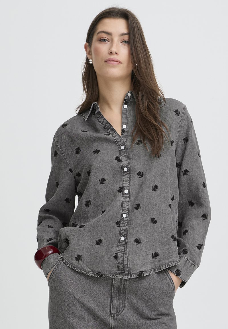 Cămașă gri din denim cu broderie florală neagră, cu nasturi în față, guler ascuțit și mâneci lungi. Purtață cu o ținută gri asortată.