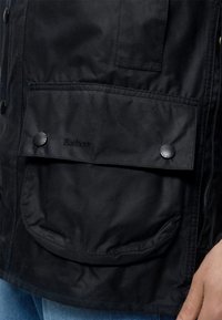 Zwarte Barbour jas zak met klep, vastgemaakt met twee zwarte drukknoopjes, een deel van een hand en blauwe spijkerbroek zichtbaar.