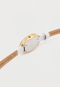 Λευκό  δερμάτινο ρολόι με χρυσή ανοξείδωτη καμπύλη, ασημένιο πίσω μέρος και κυκλικό πρόσωπο με λεπτομέρεια πλευρικού κουμπιού.