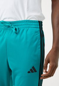 Close-up of a person wearing turquoise Adidas track pants with black stripes and logo, and a white shirt, hand relaxed by their side.