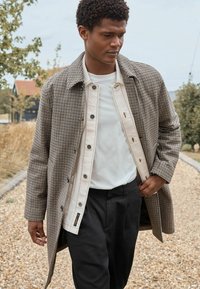 Ruskea houndstooth-takki, jossa on nappilista, suuri kaulus ja sivutaskut, kerrottu vaaleanruskean nappikauluspaidan ja valkoisen T-paidan päälle.
