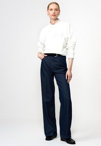 Biała bluza z ribbowanym kołnierzykiem, z logo, w połączeniu z ciemnoniebieskimi szerokimi spodniami dżinsowymi. Czarni pasek i buty dopełniają strój.