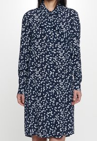 Vestito camicia blu navy con motivo a pois bianchi, dotato di colletto con fiocco, maniche lunghe e bottone frontale. Tessuto morbido e vestibilità rilassata.