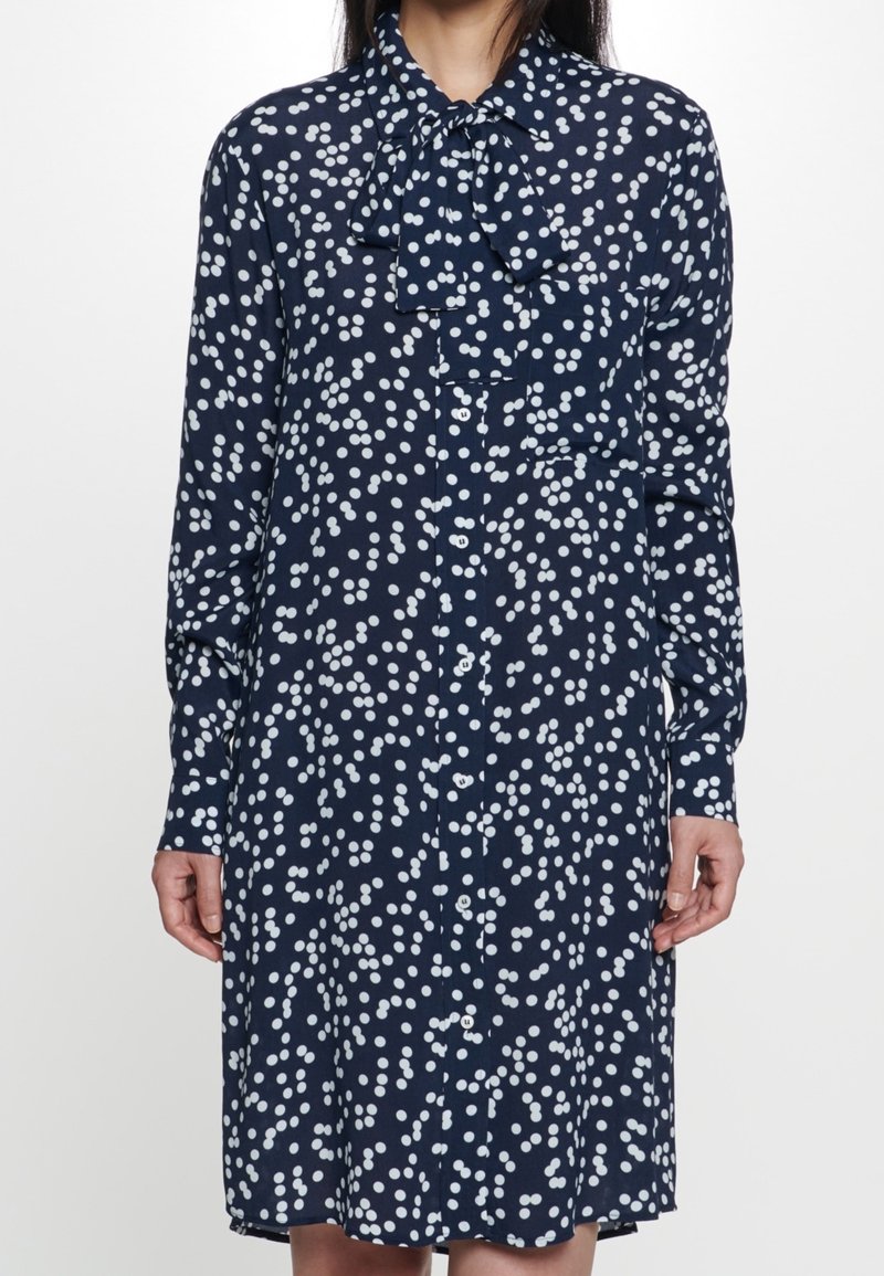 Vestito camicia blu navy con motivo a pois bianchi, dotato di colletto con fiocco, maniche lunghe e bottone frontale. Tessuto morbido e vestibilità rilassata.