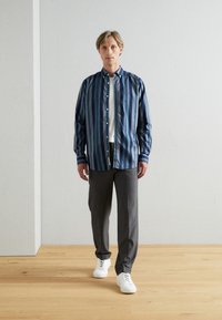 Prajčasta majica v modrih in zelenih odtenkih, zapeta, z dolgimi rokavi; sivi hlače in bele superge. Svetel leseni pod, nevtralna ozadje.