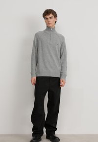 Pull en laine gris avec un col montant, fermeture éclair à l'avant et poignets côtelés, associé à un pantalon noir ample et des chaussures noires épaisses.