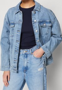 Person wearing a light blue denim jacket and jeans with a small rip on the right thigh, paired with a dark navy top.