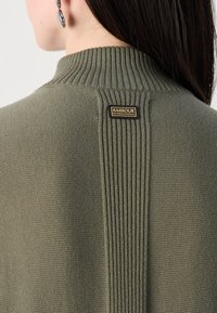 Eine Nahaufnahme des Rückens einer Person, die einen olivgrünen Pullover mit gerippten Details und einem "Barbour International" Etikett trägt.