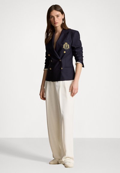 Veste de blazer double boutonnage marine avec des boutons dorés et un détail d'emblème, associée à un pantalon large crème et des chaussures de couleur claire.