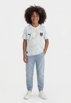 Usmievajúce sa dieťa s kučeravými vlasmi stojí oblečené v bielom futbalovom drese s vzorom, svetlomodrých džínsoch a bielych teniskách na jednoduchom pozadí.
