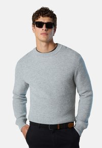 Hellgrauer, gestrickter Rundhalspullover mit strukturiertem Muster, kombiniert mit schwarzen Sonnenbrillen und einem geflochtenen Gürtel mit braunen Akzenten.