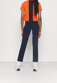 Dames sportkleding bestaat uit een oranje cropped t-shirt met een zwarte achterkant, gecombineerd met navyblauwe fitted broeken, die een gladde textuur tonen.