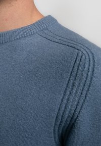 Pulover tricotat într-o nuanță de albastru deschis. Are un gât rotund și detalii texturate pe umeri. Materialul pare cald și confortabil.