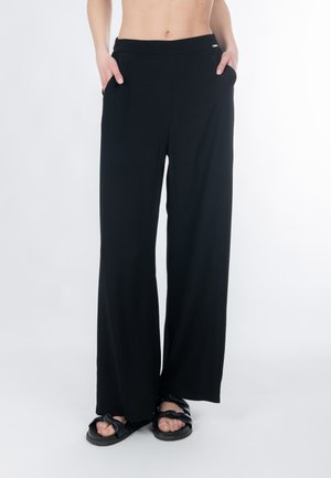 Kvinde iført løsthængende sorte bukser med hænderne i lommerne og sorte flettede sandaler, beskåret fra taljen, stående på en hvid baggrund.