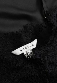 Tissu noir avec des détails en dentelle et des bords effilochés, présentant une étiquette portant le nom "NOELLA" en blanc. Texture lisse avec des motifs délicats.