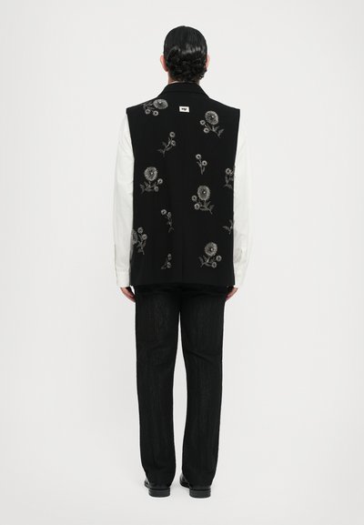 Schwarzes ärmelloses Oberteil mit floralen Stickereien, kombiniert mit einem weißen Langarmhemd und strukturierten schwarzen Hosen. Einfarbiger Hintergrund.