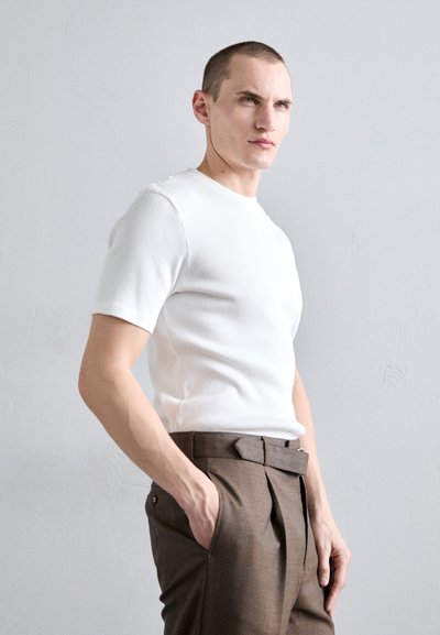 Chemise blanche à rayures, à manches courtes, associée à un pantalon marron à plis. Le mannequin se tient avec une main dans sa poche, sur un fond gris clair.