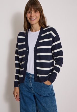 Femme souriante portant un cardigan rayé bleu marine et blanc sur une chemise blanche et un jean bleu, une main dans la poche, sur un fond uni.