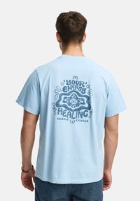 Un homme de dos portant un t-shirt bleu clair avec un graphique et un texte bleu foncé disant "Votre énergie est guérissante, inspirez expirez."