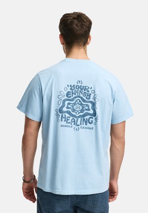 Hombre de espaldas que lleva una camiseta de color azul claro con un gráfico y texto en azul oscuro que dice "Tu energía es sanadora, inhala exhala."