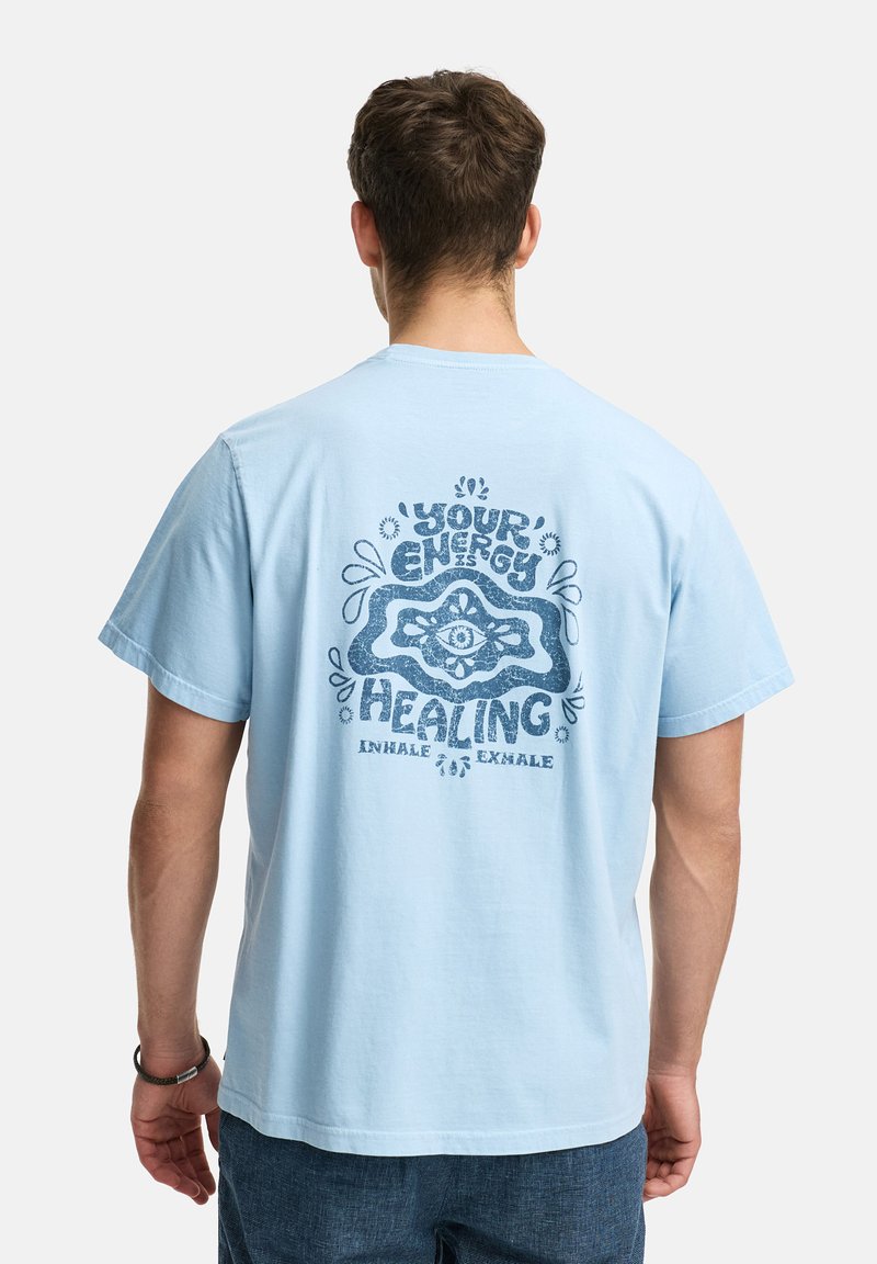 Un homme de dos portant un t-shirt bleu clair avec un graphique et un texte bleu foncé disant "Votre énergie est guérissante, inspirez expirez."