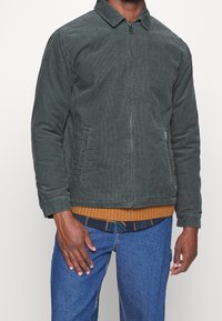 Groene corduroy jas met een kraag, voorrits en twee zijzakken. Gecombineerd met een bruine geribbelde trui daaronder.
