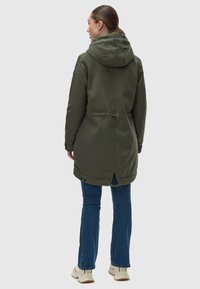 Parka verde con capucha, cintura ajustable y respiradero trasero. Combinada con jeans azules y calzado beige. Tejido suave y resistente a las inclemencias del tiempo.