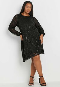 Mustapitsinen mekko, jossa on pitkät hihat, aallotettu helma ja paljettikoristelu. Kankaan pinnassa on kukkakuoseja ja rento leikkaus.