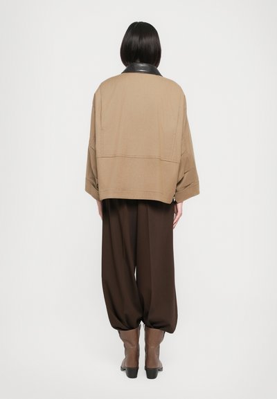 Veste beige surdimensionnée avec col en cuir noir, associée à un pantalon brun ample et des bottes ankle brunes. Tissu lisse avec un design structuré.