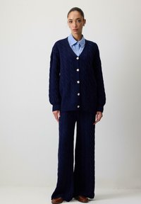 Mørkeblå kabelstrikket cardigan med store knapper, parret med matchende brede bukser. En lyseblå skjorte med krave bæres under. Glat struktur.