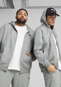 Šedé mikiny na zips s kengurovými vreckami a rebrovanými manžetami. Jedna z nich má čierne logo a je kombinovaná so šedými teplákmi a čiernym šiltovkou.