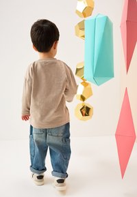 Chemise beige à manches longues, jean bleu ample et baskets blanches. Entouré de décorations géométriques en papier rose, or et bleu sarcelle.