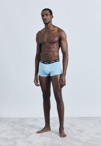 Boxer di cotone azzurro chiaro con fascia in vita nera "HUGO", caratterizzati da una vestibilità aderente e una texture liscia, indossati da un modello in piedi su una superficie grigia.