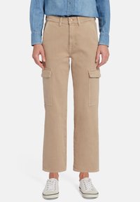 Beige cargobroek met een losse pasvorm, voorzien van twee zijzakken en een gladde textuur. Gepaard met witte sneakers en een denim shirt.