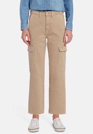 Beige cargobroek met een losse pasvorm, voorzien van twee zijzakken en een gladde textuur. Gepaard met witte sneakers en een denim shirt.