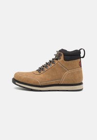 Brune suede fjellstøvler med svart tekstilkant, metalløyler, kontrasterende hvite sømmer og gummisåle. Har en pull-tab.