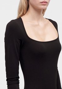 Femme portant un haut noir ajusté à manches longues avec un large décolleté carré, montrée des épaules jusqu'au milieu du torse sur un fond uni.