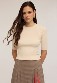 Tânără cu păr lung și ondulat, purtând cercei din aur în formă de inel, un top cu mânecă scurtă de culoare crem cu textură ribbed și o fustă cu carouri maro, cu pliseuri roșii.