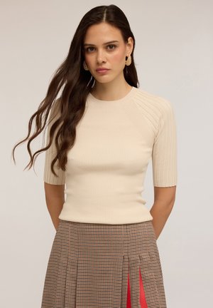 Mujer joven con cabello largo y ondulado, que lleva pendientes de aro dorados, una blusa corta de canalé color crema y una falda de cuadros marrón con pliegues rojos.