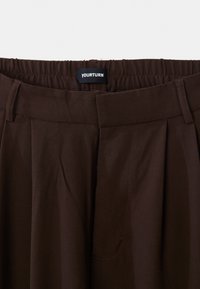 Smeđe hlače s elastičnim pojasom, petljama za remen, nabranim detaljima i prednjim zatvaranjem, sa crnom etiketom na kojoj je bijeli tekst "YOURTURN".