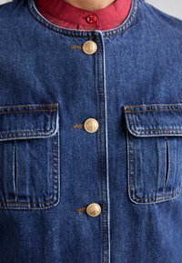 Denim jakna z oblikovano zasnovo, modro tkanino, zlatimi gumbi, dvema sprednjima žepoma in rdečim ovratnikom pod njo.