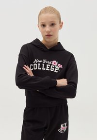 Sudadera corta negra con el texto "New York COLLEGE" y bordado floral. Combinada con pantalones negros que presentan el texto "NY COLLEGE". Tejido de algodón.