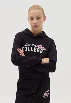 Haut à capuche noir court avec le texte "New York COLLEGE" et des broderies florales. Associé à un pantalon noir avec le texte "NY COLLEGE". Tissu en coton.