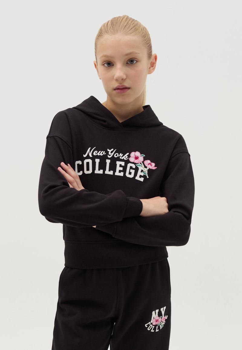 Sudadera corta negra con el texto "New York COLLEGE" y bordado floral. Combinada con pantalones negros que presentan el texto "NY COLLEGE". Tejido de algodón.