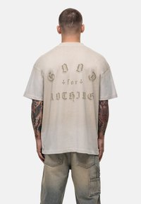 Beżowa, oversize'owa koszulka z krótkim rękawem i opadłym dekoltem, z wyblakłym napisem „BÓG za NIC” w ciemnooliwkowym kolorze, o luźnym kroju.