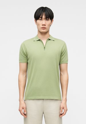 Fiatal férfi áll, világoszöld, rövid ujjú, elöl cipzáras pólóingben és bézs nadrágban, egyszínű háttér előtt.